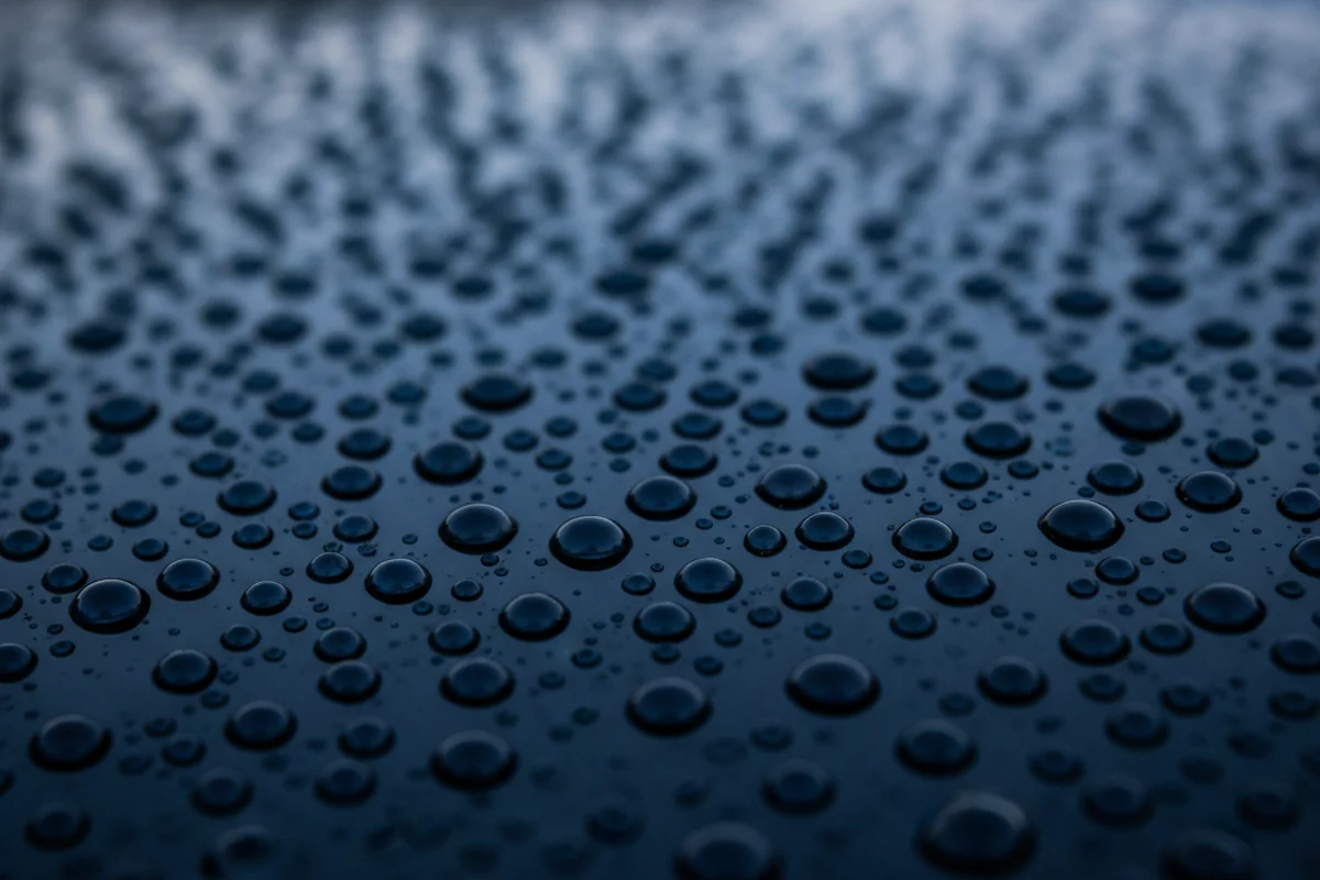 Water beads on a black ceramic-coated car hood reflecting ambient light